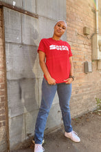 Load image into Gallery viewer, A woman wearing a red crewneck t-shirt with 'SATAN SUCKS' printed in white across the chest, paired with blue jeans and white sneakers, standing against a brick and metal wall.
